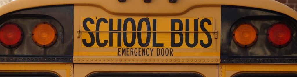 The top part of a yellow school bus with the words "School Bus" and "Emergency Door" in the middle next to a sets of red and yellow school bus lights on each side of the wording.